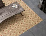 Un suelo hidráulico con diseño geométrico en tonos tierra y ocre, junto a un suelo de cemento pulido. Una mesa de madera rústica y una silla completan la escena.