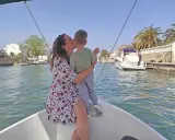 Madre besa a su hijo en un paseo en barco por los canales de Empuriabrava, con palmeras y casas blancas al fondo.
