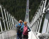 Para idzie przez długi most wiszący w Czechach, robiąc zdjęcia. W tle widać las i innych turystów.