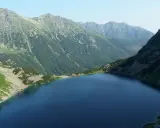 Górski krajobraz z jeziorem i stromymi zboczami. W oddali widać pasmo górskie, które przypomina najwyższy szczyt w Polsce.