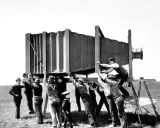 Grupa mężczyzn w historycznych strojach ustawia gigantyczny aparat fotograficzny. Wyobraź sobie, kiedy wynaleziono aparat fotograficzny i jak wielkie musiały być pierwsze urządzenia!