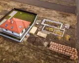 Budowa domu na działce rolnej. Widać fundamenty, sterty drewna i cegieł, gotowy dom. To może być początek planowania, jak podzielić działkę rolną.