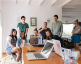 Grupa studentów na którym roku studiów są praktyki, pochylona nad laptopami, dyskutuje przy drewnianym stole.