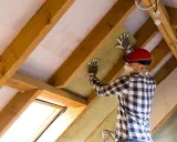 Uomo con mascherina e guanti installa materiale isolante tra le travi di legno di un sottotetto. È chiaro che conviene isolare il sottotetto per migliorare il comfort.