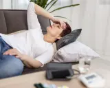 Donna sdraiata sul divano con la mano sulla fronte, visibilmente affaticata e con la paura di svenire. Sul tavolino, un misuratore di pressione.