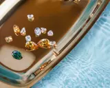 Un singolo zaffiro accento verde smeraldo risalta tra diamanti e pietre color miele su un vassoio dorato, riflettendo la luce.