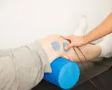 Rehabilitacja kolana w domu: elektrody na skórze, wałek do masażu pod nogą, ręka terapeuty.
