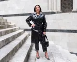 Donna con un audace outfit rock: trench in ecopelle nera, jeans scuri, décolleté borchiate e borsa a tracolla.