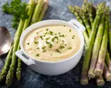 Un bol de sauce mousseline onctueuse, garni de persil frais, est présenté avec des asperges vertes et une cuillère.