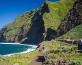 Grüne Berge und rote Felsen prägen die Küste der Madeira Inselgruppe. Ein Weg führt zu kleinen Häusern.
