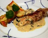 En rullad kyckling insvept i bacon, serverad med en krämig sås till kyckling och gratinerade potatisbitar med gröna blad.