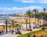 En livlig strandpromenad i Sitges, Spanien, med palmer, en sandstrand, båtar i hamnen och människor som njuter av solen.