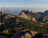 Edificio con antena en la cima de una montaña rocosa, con vistas a un paisaje montañoso y brumoso que se extiende hasta el horizonte, evocando la sensación de estar en planos cerca de Barcelona.