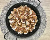 Almejas con jamón en una paellera, con trocitos de jamón y perejil picado, sobre un mantel de madera.