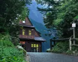 Drewniane schronisko turystyczne w otoczeniu zieleni, z kamienną drogą prowadzącą do wejścia. W tle widać górzysty krajobraz.
