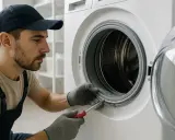 Técnico con gorra y overol trabaja en la lavadora, usando un destornillador para arreglar la goma. Se puede cambiar la goma de la lavadora.