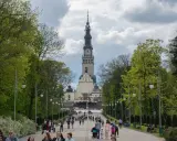 Aleja prowadząca do imponującej wieży klasztornej, jednego z ważnych sanktuariów w Polsce. Ludzie spacerują, podziwiając architekturę i otaczającą zieleń.