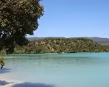 Vue paisible de l'étang de la Bonde, avec ses eaux turquoise bordées de verdure et de collines boisées sous un ciel bleu clair.
