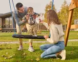 Radosne zabawy w ogrodzie: tata podtrzymuje dziecko na huśtawce, mama zachęca do zabawy.
