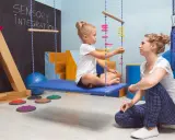 Dziecko siedzi na huśtawce w sali integracji sensorycznej, obok siedzi terapeuta.