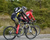 Dwóch kolarzy górskich w kaskach i strojach sportowych jedzie po szutrowej drodze. Jeden z nich, z tyłu, ma na nogawkach napis "Scott". Słychać, jak rower skrzypi podczas pedałowania.
