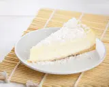 Une part de tarte noix de coco, crémeuse et généreusement saupoudrée de noix de coco râpée, repose sur une assiette blanche, prête à être dégustée.