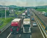 Ciężarówki na autostradzie, jedna czerwona Scania wyprzedza białą. W tle inne pojazdy i krajobraz z pagórkami.