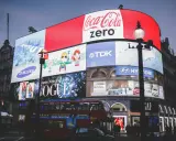 Wielkie ekrany reklamowe na placu, prezentujące różne rodzaje reklamy: Coca-Cola Zero, Samsung, Vogue, Hyundai, TDK. Widać też autobus i ludzi.