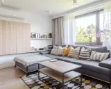 Przytulny salon w stylu dom kostka. Szara kanapa z poduszkami, drewniany stolik kawowy, jasne meble na ścianie i duża sofa.