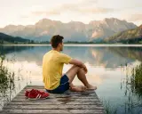 Mann in gelbem Shirt sitzt auf Steg am See, blickt auf Berge. Seine roten Schuhe liegen daneben. Perfekt für die taper week.