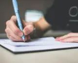 Dłoń pisze niebieskim długopisem w notatniku. To może być początek pisania opinii, esej przykład.