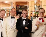 Four men in stylish suits and bow ties, embodying creative black tie, pose at a bar. One holds a cigar, another a drink.