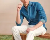 A man poses in a blue denim shirt and crisp white jeans, looking relaxed on a wooden bench.
