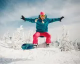 Dziewczyna w różowej czapce i goglach skacze na snowboardzie na tle zaśnieżonych drzew. To jest właśnie snowboard!