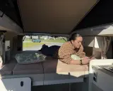 Une jeune femme se détend sur le lit de son camping-car j80 interieur, regardant son téléphone. Dehors, le soleil brille.