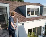 Junge Person auf Balkon blickt zu Frau in Schleppgaube. Verschiedene Dachgauben Arten sind zu sehen.