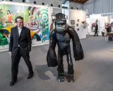 Mann in Anzug neben Gorilla-Skulptur auf Kunstmesse. Bunte Gemälde im Hintergrund zeigen aktuelle Kunst.
