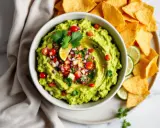 Un bol de guacamole frais, garni de tomates, oignons rouges et coriandre, prêt à être dégusté avec des chips tortilla. La meilleure recette guacamole !
