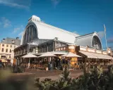 Människor sitter vid bord utomhus framför den imponerande **stora saluhallen** under en klarblå himmel.