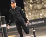 Kobieta w czapce z daszkiem i sportowym stroju rozciąga nogę na tle miejskiego krajobrazu.