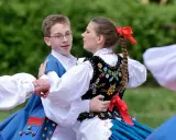 Młoda para w strojach ludowych tańczy mazura. Chłopiec w niebieskiej kamizelce, dziewczyna w haftowanej kamizelce i warkoczu z czerwoną wstążką.