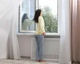 Mujer abre ventana para ventilar la casa en invierno. Reflejo en el cristal muestra a otra persona.