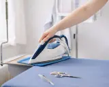 Eine Hand hält ein Bügeleisen auf einem Bügelbrett. Neben dem Bügeleisen liegen eine Schere und ein Federkiel. Der leinen trockner ist bereit für die Wäsche.