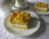 Sernik z brzoskwiniami na białym talerzyku, z kruszonką na wierzchu. Obok filiżanka i dzbanek.