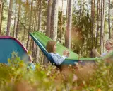 Två kvinnor njuter av en lugn stund i en hängmatta i skogen, med ett tält i närheten. En perfekt bild av campa Sverige.