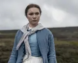 Florence Pugh i en blå klänning och jacka, med en scarf runt halsen, står i ett öppet landskap. En scen från filmer med Florence Pugh.