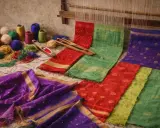 Vibrant Indian textiles, including colorful yarn balls, a weaving shuttle, and richly patterned fabrics in red, green, and purple, are laid out near a loom.
