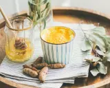 Aromaticzne złote mleko w kubeczku, obok słoiczek z miodem i korzenie kurkumy. Idealne na chłodne wieczory.