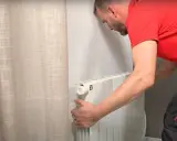 Un homme en t-shirt rouge et pantalon de travail ajuste un radiateur électrique blanc, prêt pour son branchement.