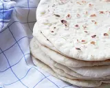 En hög med nybakade tortillabröd, perfekta för ditt nästa tortilla recept. Bredvid ligger en blåvitrutig kökshandduk.